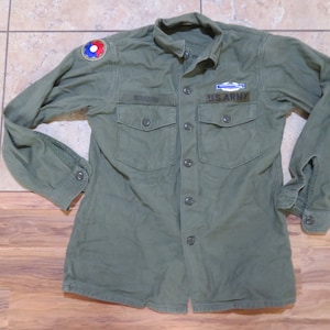 Puede incluir: Una camisa militar verde de manga larga con cierre de botones. La camisa tiene dos bolsillos en el pecho y un parche en la manga izquierda que dice "U.S. ARMY".