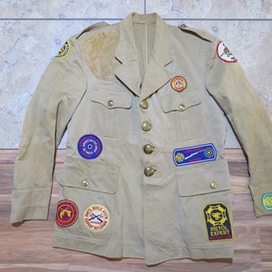 Peut inclure: Veste beige ornée de nombreux écussons, dont "American Rifle Assn" et "Pistol Expert". La veste a une fermeture boutonnée, quatre poches et des épaulettes. Les écussons présentent du texte et divers emblèmes.