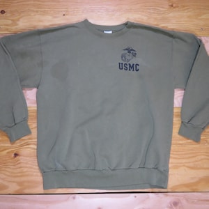 May include: Olive green sweatshirt with the text "USMC" and an emblem printed on the upper chest. The long-sleeved crew neck top is made of a soft material. The sweatshirt is laid flat on a wooden surface.