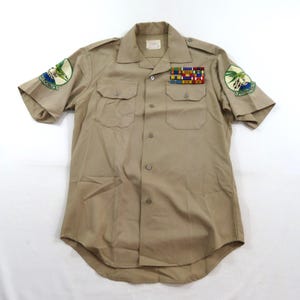 May include: A tan short-sleeved button-down shirt featuring two chest pockets, shoulder epaulets, and embroidered circular patches on both sleeves. A multi-colored ribbon bar is pinned above the left chest pocket, and a label is inside the collar.