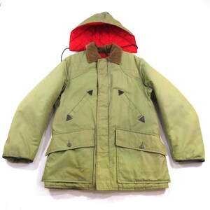Puede incluir: Una chaqueta de color verde oliva con cuello de pana marrón, capucha desmontable con forro acolchado rojo brillante, dos bolsillos en el pecho con detalles triangulares de cuero y dos bolsillos grandes con solapa y botón al frente.