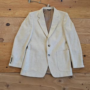 Könnte beinhalten: Ein cremefarbener Blazer mit einem gesprenkelten Muster. Der Blazer hat zwei Knopfverschlüsse und zwei Eingrifftaschen.