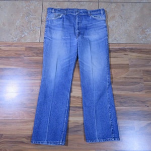 May include: A pair of light blue denim jeans with a classic five-pocket design. The jeans have a faded wash and a relaxed fit, with a straight leg that widens slightly towards the bottom. The jeans are laid flat on a wooden surface.