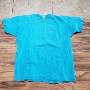 Könnte beinhalten: Ein türkisblaues T-Shirt mit Rundhalsausschnitt und einer Tasche auf der linken Brust. Das kurzärmelige Shirt ist aus einem weichen Material gefertigt und liegt flach auf einer Holzoberfläche.