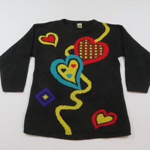 May include: Black long-sleeve sweater with colorful heart and geometric designs. The sweater features a large red heart with a checkered pattern, a blue heart with yellow accents, and a yellow wavy line. Additional hearts and a diamond shape are also present.