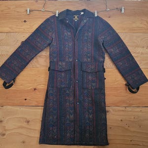 May include: A long, patterned coat in shades of blue, burgundy, and tan, with a collar and two front pockets. The coat is hanging on a wooden surface. The coat has a button-up front and adjustable straps at the cuffs.