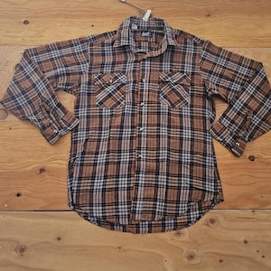 Puede incluir: Una camisa de franela a cuadros marrón, negra y azul con mangas largas y botones. La camisa tiene dos bolsillos en el pecho y un cuello. Se muestra sobre una superficie de madera.