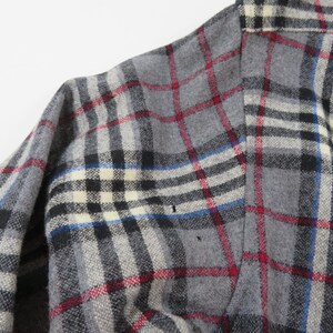 May include: A long-sleeved shirt featuring a grey, black, white, and red plaid pattern. The fabric has a textured appearance with a collar visible at the top. The garment displays a classic button-down construction with a standard shoulder seam.