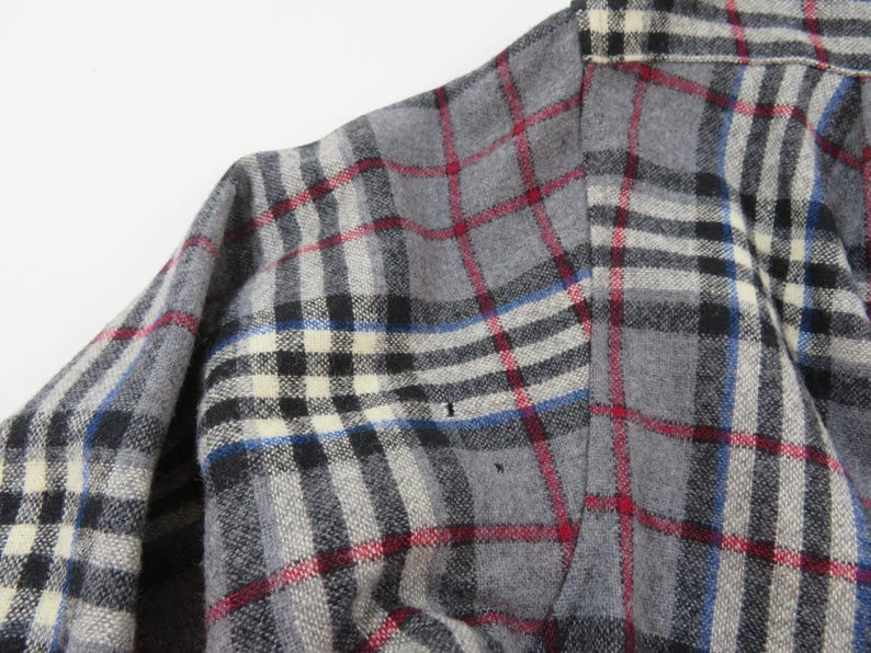 May include: A long-sleeved shirt featuring a grey, black, white, and red plaid pattern. The fabric has a textured appearance with a collar visible at the top. The garment displays a classic button-down construction with a standard shoulder seam.