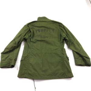 May include: An olive green field jacket featuring long sleeves, a high collar, and two front pockets. The back of the jacket displays the black stenciled text "SUPPLY" above the number "0042". The garment includes shoulder epaulets and a drawstring hem.