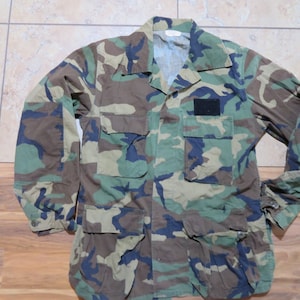 Könnte beinhalten: Tarnmusterjacke mit mehreren Taschen und Knopfleiste. Die Jacke weist eine Mischung aus Grün-, Braun- und Schwarztönen auf, mit einem kleinen schwarzen Aufnäher auf der Brust. Die Ärmel haben geknöpfte Manschetten.