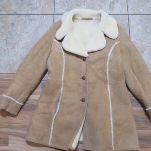 Op de afbeelding: Een bruin suède jas met een crème shearling voering en kraag. De jas heeft een knoopsluiting en witte biezen. De jas is een middellange stijl.