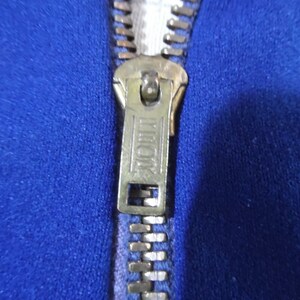 May include: A vibrant royal blue fabric garment featuring a metallic brass-toned zipper closure. The zipper pull tab is rectangular with the word TALON embossed in a clear, capitalized font, centered between two horizontal lines on the metal surface.