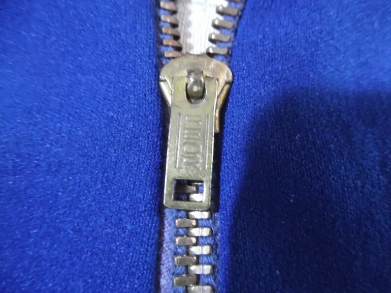 May include: A vibrant royal blue fabric garment featuring a metallic brass-toned zipper closure. The zipper pull tab is rectangular with the word TALON embossed in a clear, capitalized font, centered between two horizontal lines on the metal surface.