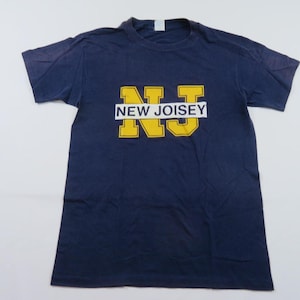 May include: Navy blue t-shirt with the text "NEW JOISEY" in yellow block letters. The text is centered on the front of the shirt. The shirt has short sleeves and a crew neck.