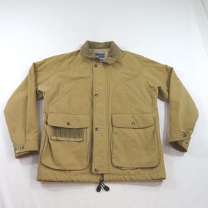 May include: A tan field jacket featuring a corduroy collar, a front button placket, and two large lower pockets with flaps. The right pocket includes an external elasticised shell holder. The jacket has long sleeves with buttoned cuffs and a drawstring hem.