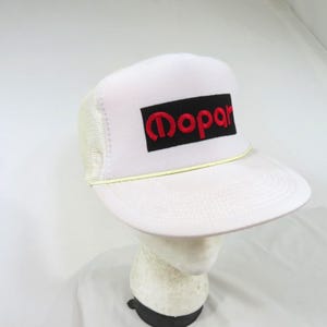 Puede incluir: Una gorra de camionero blanca con un parche rectangular negro en el panel frontal que tiene la palabra Mopar bordada en letras rojas. La gorra incluye una parte trasera de malla blanca, un panel frontal blanco sólido y un cordón amarillo fino.