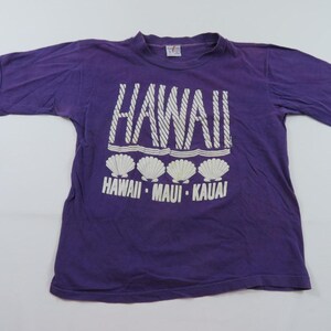 Puede incluir: Una camiseta morada con texto y gráficos blancos. La palabra "HAWAII" está impresa en letras grandes y estilizadas sobre cuatro gráficos de conchas marinas. Debajo de las conchas, el texto dice "HAWAII - MAUI - KAUAI".