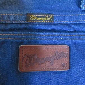 May include: Close-up of a pair of blue denim jeans with a brown leather Wrangler label. The label has the word "Wrangler" embossed in the leather.