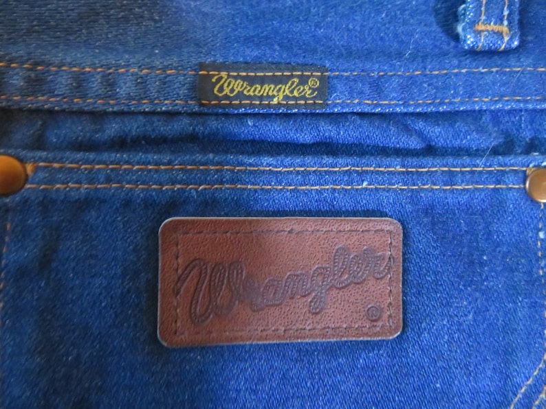 May include: Close-up of a pair of blue denim jeans with a brown leather Wrangler label. The label has the word "Wrangler" embossed in the leather.