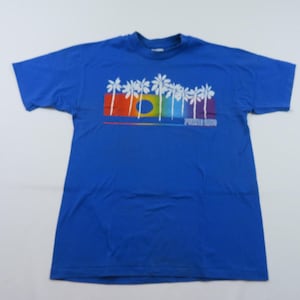 May include: A royal blue t-shirt featuring a colorful graphic of white palm trees, a rainbow, and the text "Florida". The design includes red, orange, yellow, green, and purple hues. The shirt is made of a soft material.