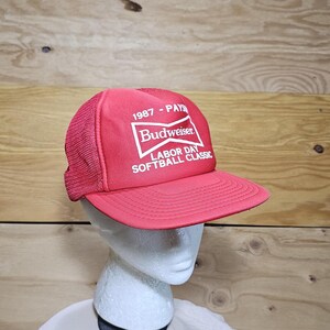 Op de afbeelding: Rode Budweiser baseballpet met witte tekst en logo. De pet heeft de tekst "1987 - PAYS" en "Labor Day Softball Classic". De pet heeft een mesh achterkant en een gebogen klep.