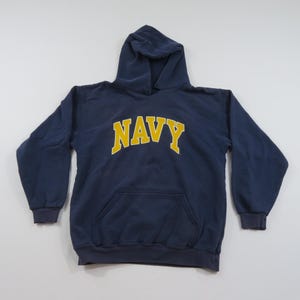 Könnte beinhalten: Ein marineblauer Kapuzenpullover mit dem Wort "NAVY" in gelben Blockbuchstaben auf der Brust. Der Pullover hat eine Vordertasche und gerippte Bündchen und Saum. Die Kapuze ist ungefüttert.