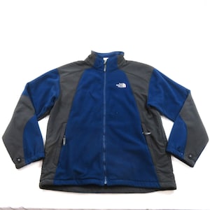 May include: A long-sleeve fleece jacket featuring a color-blocked design in dark blue and charcoal gray. The jacket has a full-length front zipper, a stand-up collar, and the white The North Face logo embroidered on the upper left chest area.