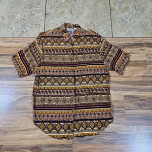 Puede incluir: Camisa de manga corta con botones y estampado geométrico en tonos marrón, beige, burdeos y dorado. La camisa presenta un cuello y un corte clásico, adecuada para ropa informal. El patrón incluye diseños de diamantes y rayas.