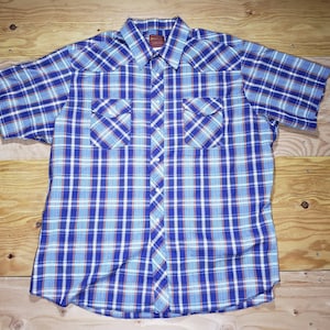 Puede incluir: Camisa de manga corta a cuadros azules, blancos y rojos. Tiene dos bolsillos en el pecho y cierre de botones. La tela parece ligera, ideal para el verano. El diseño a cuadros es clásico.