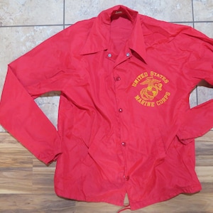 Op de afbeelding: Een felrode nylon jas met lange mouwen en een kraag. De jas heeft een knoopsluiting aan de voorkant en een trekkoord aan de onderkant. De woorden "United States Marine Corps" zijn in goud op de borst geborduurd.
