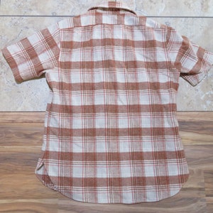 May include: Short-sleeved, button-down shirt with a plaid pattern in shades of brown, white, and red. The shirt has a classic collar and a curved hemline. The fabric appears to be a soft, woven material.