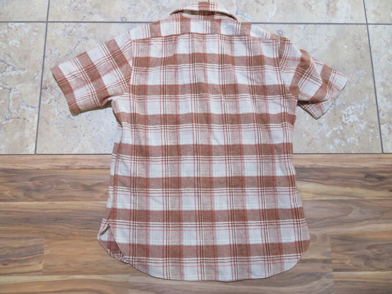 May include: Short-sleeved, button-down shirt with a plaid pattern in shades of brown, white, and red. The shirt has a classic collar and a curved hemline. The fabric appears to be a soft, woven material.