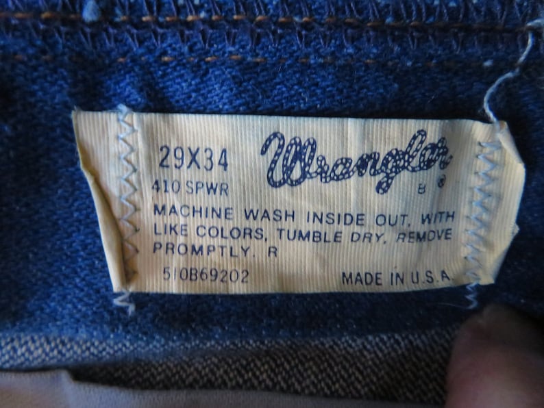 May include: A white Wrangler jeans care label with black text. The label reads "29X34", "410 SPWR", "MACHINE WASH INSIDE OUT, WITH LIKE COLORS, TUMBLE DRY, REMOVE PROMPTLY. R", "510B69202", and "MADE IN U.S.A."