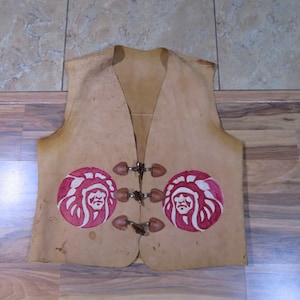 Pode incluir: Colete de couro castanho com decote em V e três fechos de alternância. O colete apresenta dois desenhos circulares vermelhos e brancos com a cabeça de um chefe nativo americano. O colete é de estilo vintage.