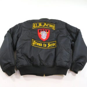 May include: A black bomber jacket featuring a large embroidered patch on the back. The patch displays a red shield with a gray fist holding a lightning bolt, topped with the yellow text U.S. Army and the yellow text Proud to Serve below it.
