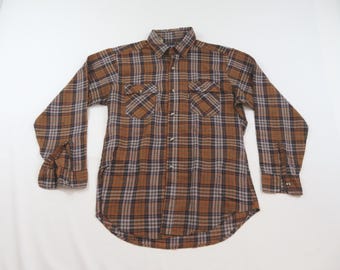 May include: A long-sleeve button-down shirt featuring a brown, tan, and dark gray plaid pattern. The shirt includes a standard collar, a full-length front button closure, and two chest pockets with buttoned flaps. The cuffs also feature buttons.