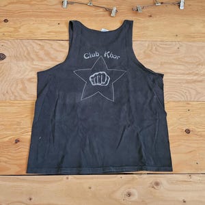 Puede incluir: Una camiseta sin mangas gris oscuro con el texto "Club Khor" encima de una estrella blanca con un puño cerrado. La camiseta sin mangas está hecha de un material suave y está colocada sobre una superficie de madera.