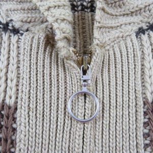 Puede incluir: Primer plano de un su&eacute;ter de punto beige y marr&oacute;n con cremallera. El su&eacute;ter presenta un patr&oacute;n texturizado con dise&ntilde;os verticales y geom&eacute;tricos. La cremallera tiene un tirador plateado con un asa circular.