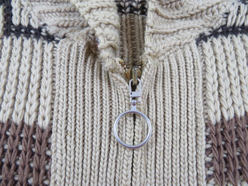 Puede incluir: Primer plano de un su&eacute;ter de punto beige y marr&oacute;n con cremallera. El su&eacute;ter presenta un patr&oacute;n texturizado con dise&ntilde;os verticales y geom&eacute;tricos. La cremallera tiene un tirador plateado con un asa circular.