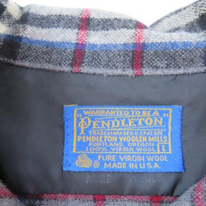 May include: A grey, black, white, blue, and red plaid wool garment featuring a blue rectangular label with yellow text that reads: WARRANTED TO BE A PENDLETON, PENDLETON WOOLEN MILLS, PORTLAND, OREGON, 100% VIRGIN WOOL, L, PURE VIRGIN WOOL, MADE IN U.S.A.
