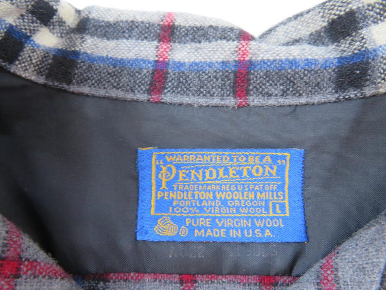 May include: A grey, black, white, blue, and red plaid wool garment featuring a blue rectangular label with yellow text that reads: WARRANTED TO BE A PENDLETON, PENDLETON WOOLEN MILLS, PORTLAND, OREGON, 100% VIRGIN WOOL, L, PURE VIRGIN WOOL, MADE IN U.S.A.