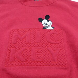Puede incluir: Una sudadera roja con un dise&ntilde;o "MICKEY" texturizado en un tono m&aacute;s oscuro de rojo. Encima del texto hay una peque&ntilde;a imagen bordada de Mickey Mouse. La sudadera tiene cuello redondo y est&aacute; hecha de un material suave.