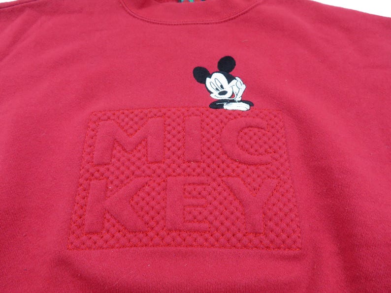 Puede incluir: Una sudadera roja con un dise&ntilde;o "MICKEY" texturizado en un tono m&aacute;s oscuro de rojo. Encima del texto hay una peque&ntilde;a imagen bordada de Mickey Mouse. La sudadera tiene cuello redondo y est&aacute; hecha de un material suave.