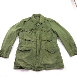 May include: An olive green field jacket featuring a pointed collar, long sleeves with buttoned cuffs, and four large front pockets with button-down flaps. The jacket includes shoulder epaulets and a central zipper closure with a protective storm flap.