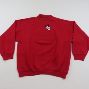 Puede incluir: Una sudadera roja con cuello alto y mangas largas. La sudadera presenta un dise&ntilde;o bordado de Mickey Mouse en la parte superior izquierda del pecho y las palabras "Mickey" en relieve debajo. Los pu&ntilde;os y el dobladillo son acanalados.