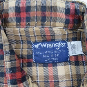 May include: A long-sleeve button-down shirt featuring a plaid pattern in tan, black, and red. A blue Wrangler brand label is stitched to the inside collar, displaying the text Wrangler, Single Needle Tailoring, 14 1/2 x 32, Long Tails, Made in U.S.A.