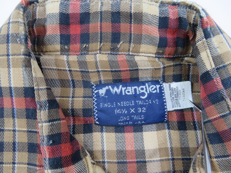 May include: A long-sleeve button-down shirt featuring a plaid pattern in tan, black, and red. A blue Wrangler brand label is stitched to the inside collar, displaying the text Wrangler, Single Needle Tailoring, 14 1/2 x 32, Long Tails, Made in U.S.A.
