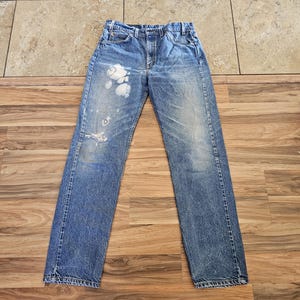 May include: Blue denim jeans with distressed details, including a ripped knee and white paint splatters. The jeans are laid flat on a wooden surface, showcasing their vintage style. A classic, casual apparel item.