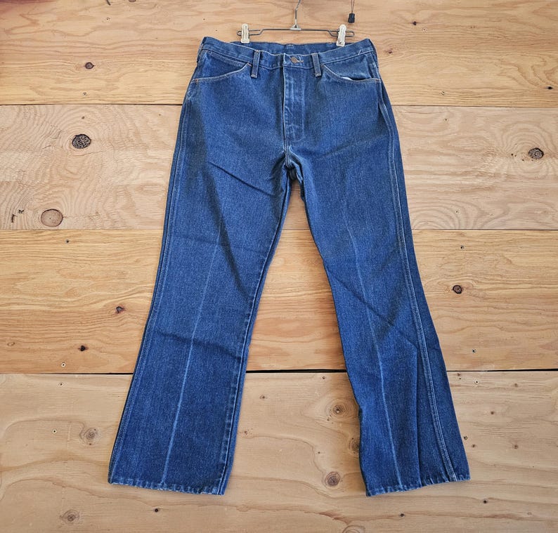 May include: Blue denim jeans with a high waist and flared leg design. The jeans are hanging on a metal hanger against a light brown wooden background. The jeans have a classic five-pocket design.